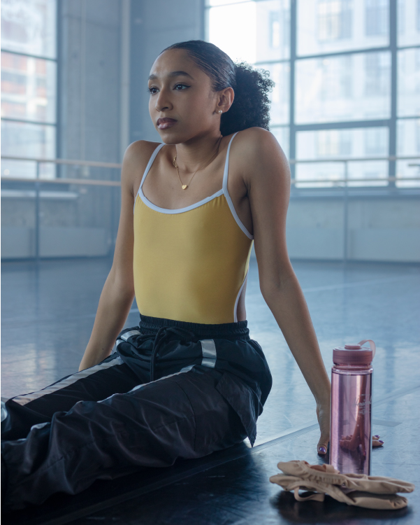 eine Frau, die in einem Ballettstudio sitzt, mit Ballettschuhen und einer rosa Herschel-Wasserflasche neben sich.