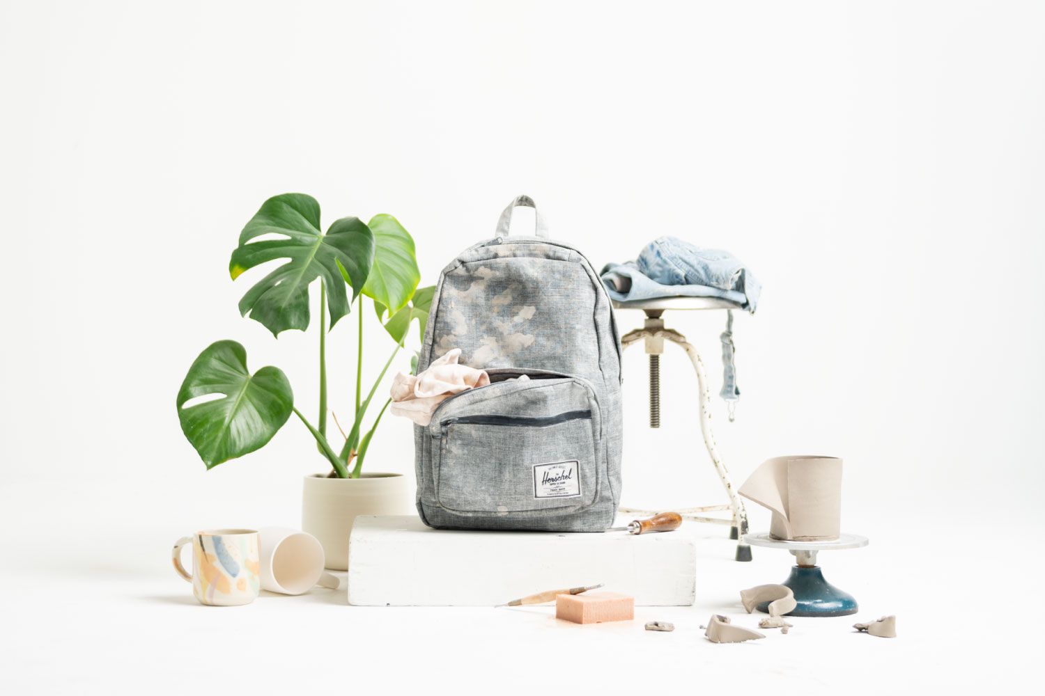 A Herschel Classic Backpack XL in Raven Crosshatch next to a potted plant and a potter stool, apron, and some pottery tools