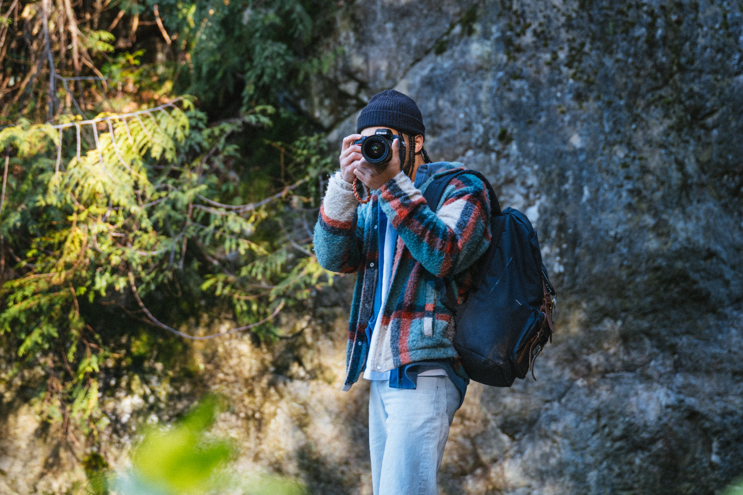 A person in the woods wearing their Herschel Little America Backpack while holding up their camera taking a photo.