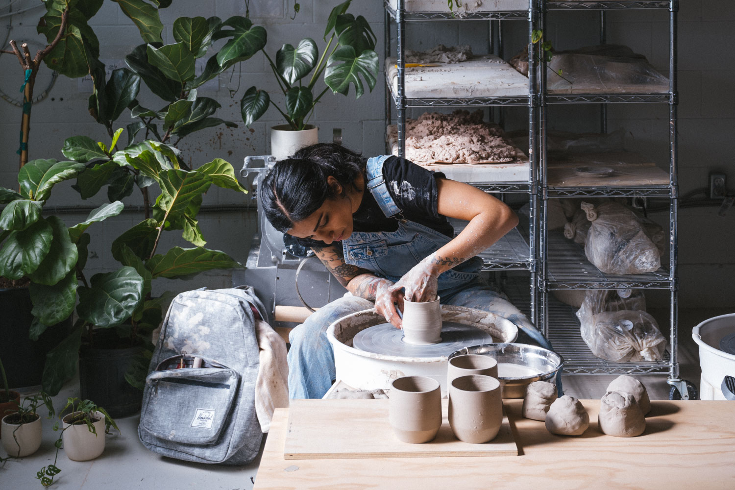 A person in a pottery studio making a cup on the pottery wheel with their Herschel Classic Backpack XL on the ground behind them with their tools sticking out.
