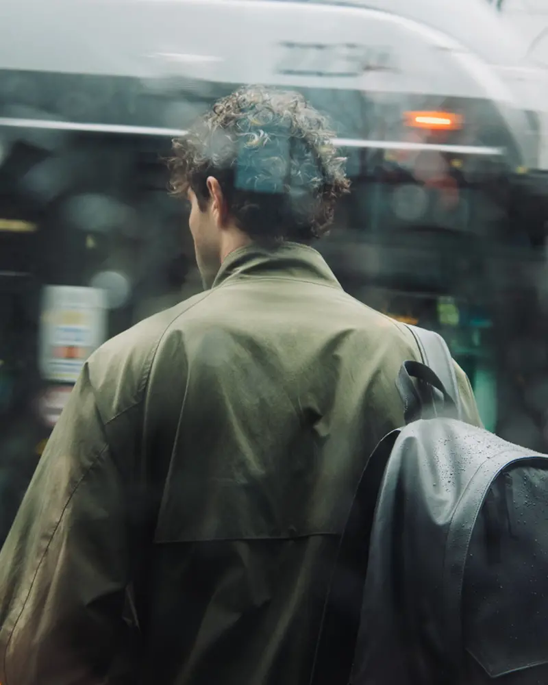 An image of a man standing in the rain, waiting for a bus, wearing a black Herschel Alberni Weather Resistant Backpack 